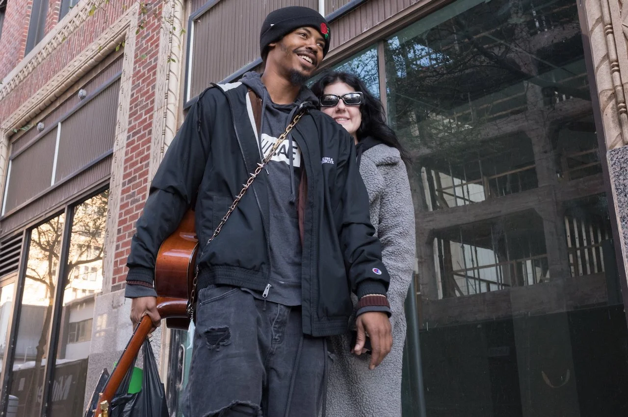 A young man and woman smiling outdoors on a city street. The man is wearing a black hoodie, black jacket, black beanie, and sunglasses, carrying a guitar on his back. The woman is wearing a gray fuzzy coat and sunglasses. They are standing in front o