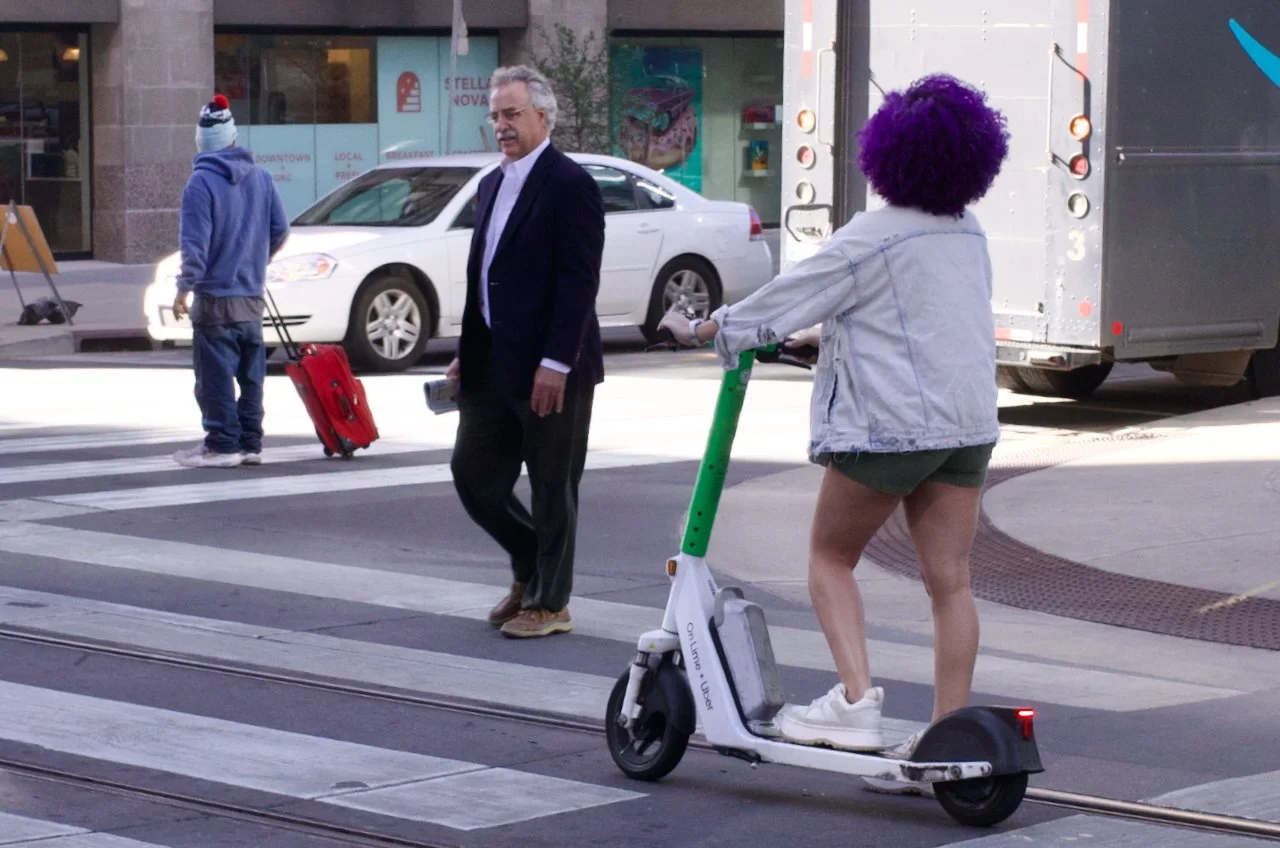 A woman with purple hair riding an electric scooter across the street while a man in a suit waits to cross, and a person with a blue hoodie and red suitcase walks in the background.