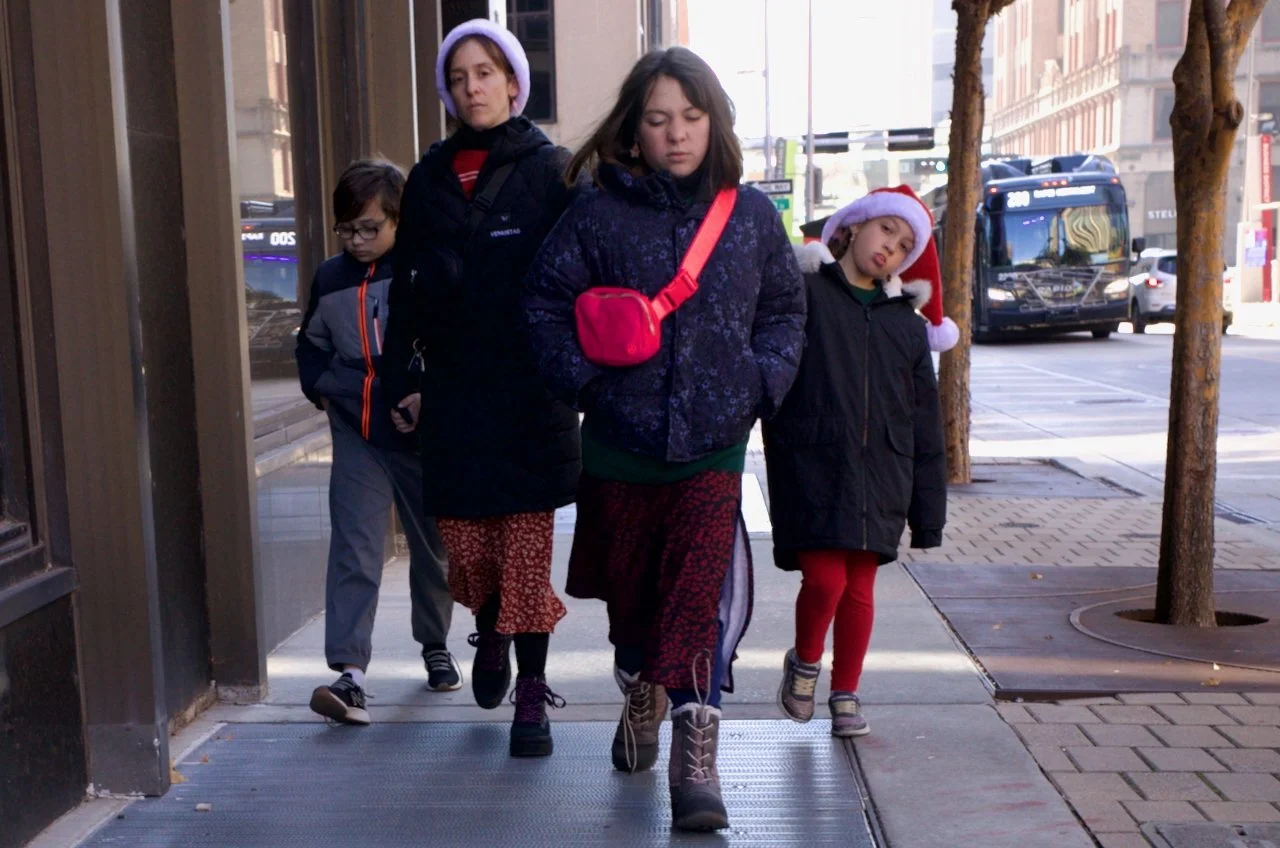Four children walking on a city sidewalk, with two wearing Santa hats, one with a red purse, and all dressed warmly.