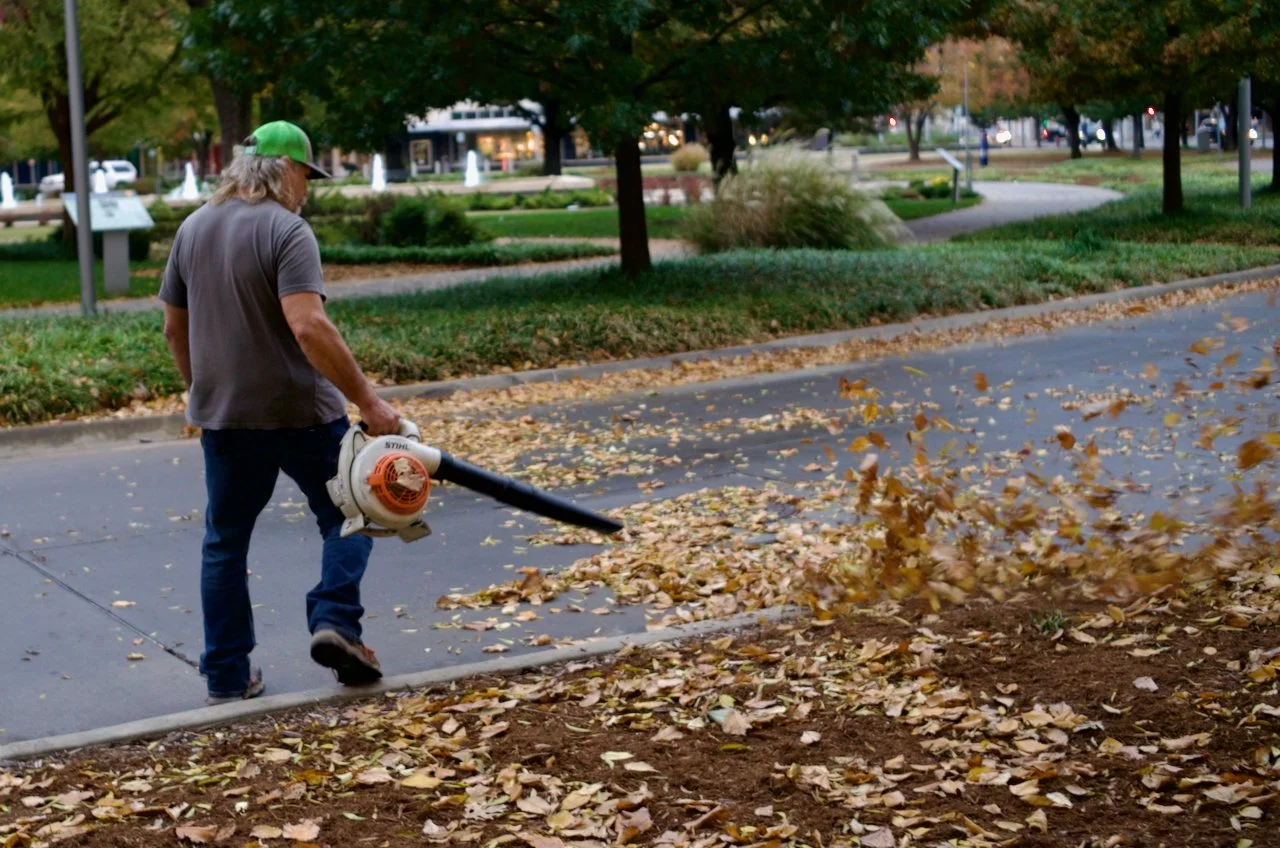 A man in a green cap and gray t-shirt uses a leaf blower to clear fallen leaves from the sidewalk in a park with trees and green grass.