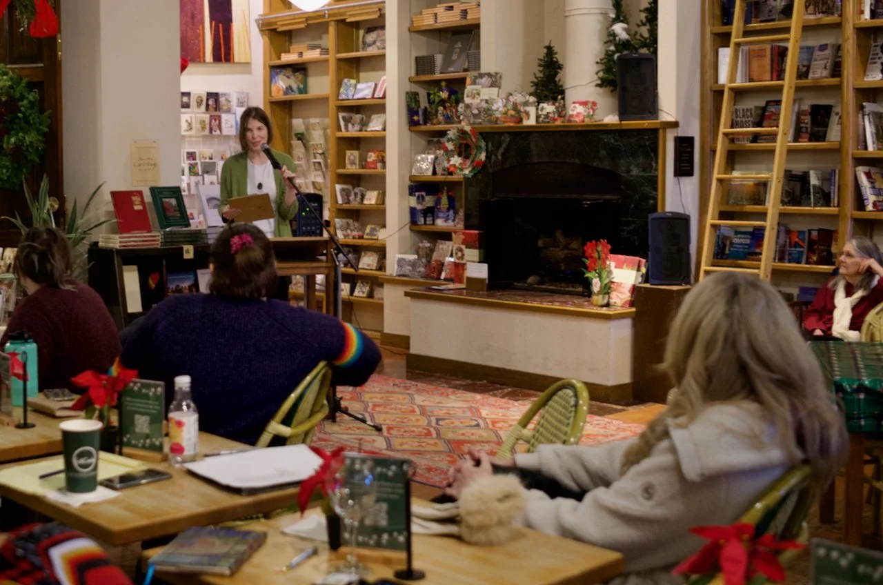 A woman at OKC Full Circle Bookstore's poetry night stands at a podium with a microphone, reading or speaking to an audience in a cozy bookstore or cafe decorated for Christmas, with bookshelves, holiday decorations, and people seated at tables.