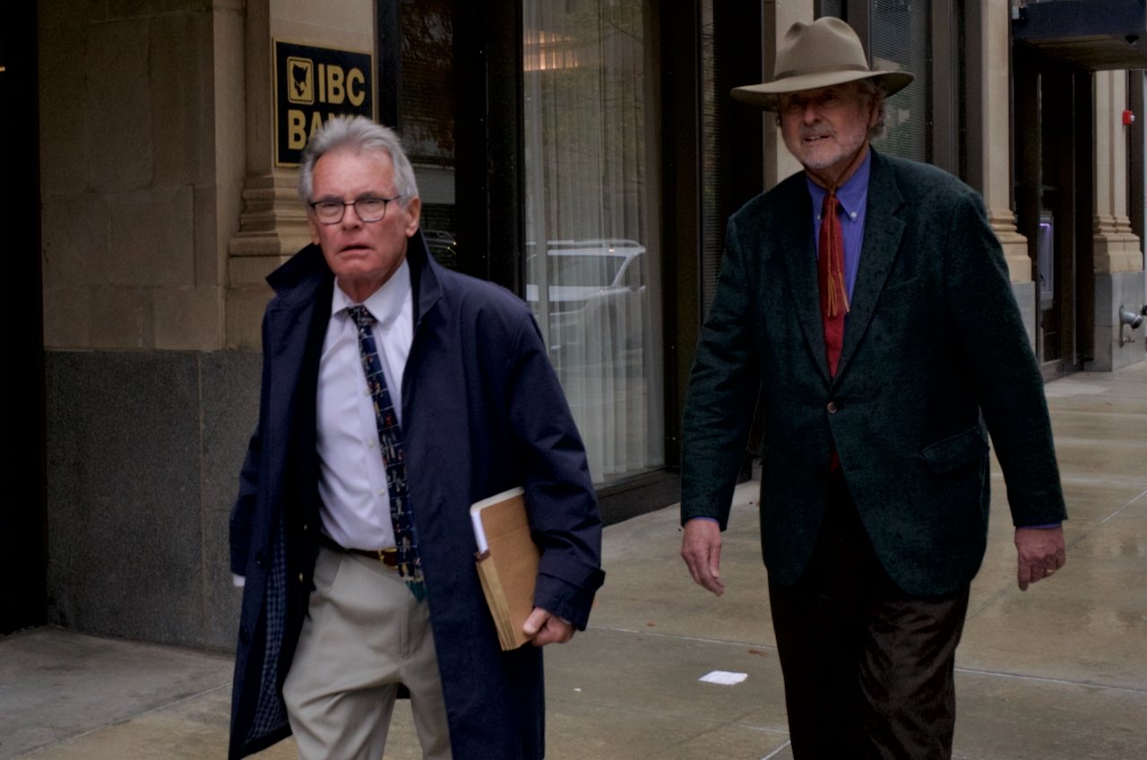 Two older men walking on the sidewalk outside a building with a sign that says 'IBC Bank.' One man is wearing a navy jacket and tie, glasses, and holding a folder or notebook. The other man is wearing a green blazer, a wide-brimmed hat, and a blue sh