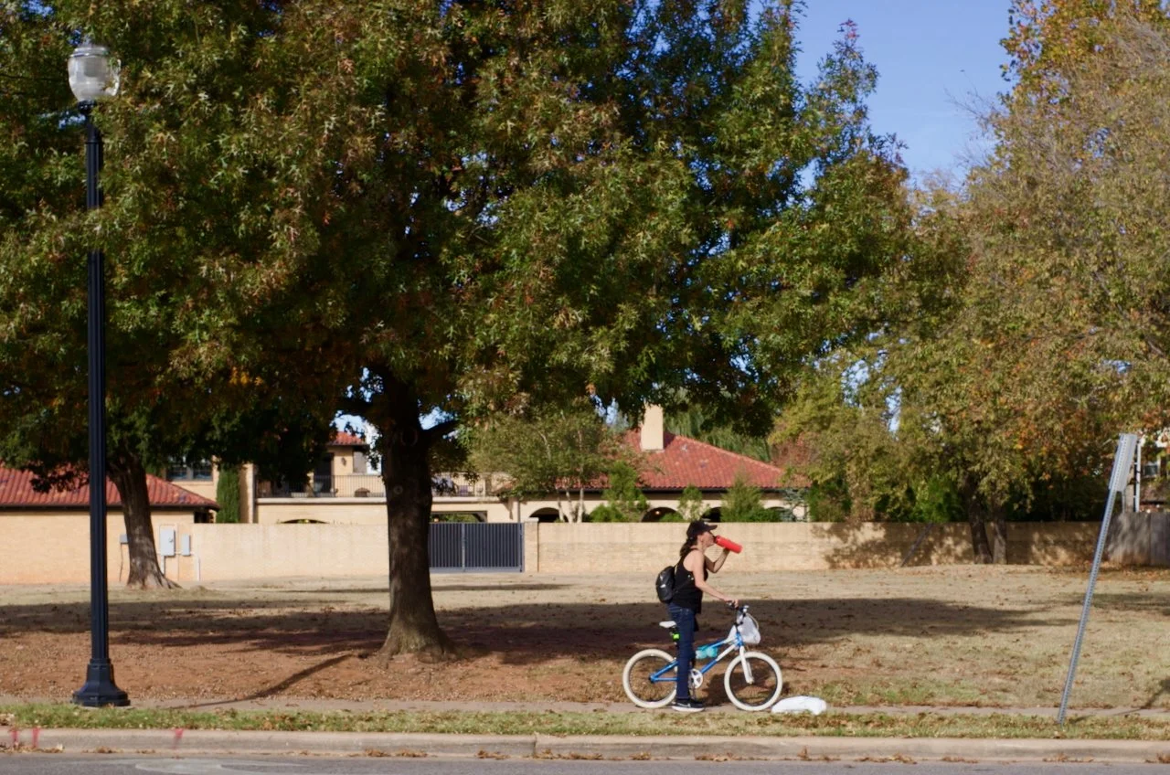 A girl riding a blue and white bicycle, drinking from a red water bottle, on the sidewalk in front of trees and a park area during daytime.