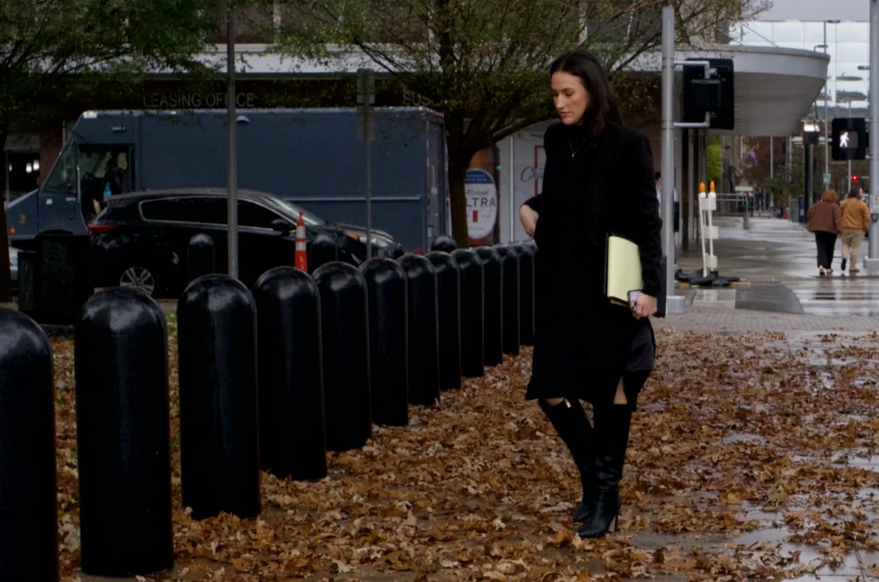 A woman with dark hair dressed in all black, holding a yellow folder, walking along a leaf-covered sidewalk in an urban area on a cloudy day.