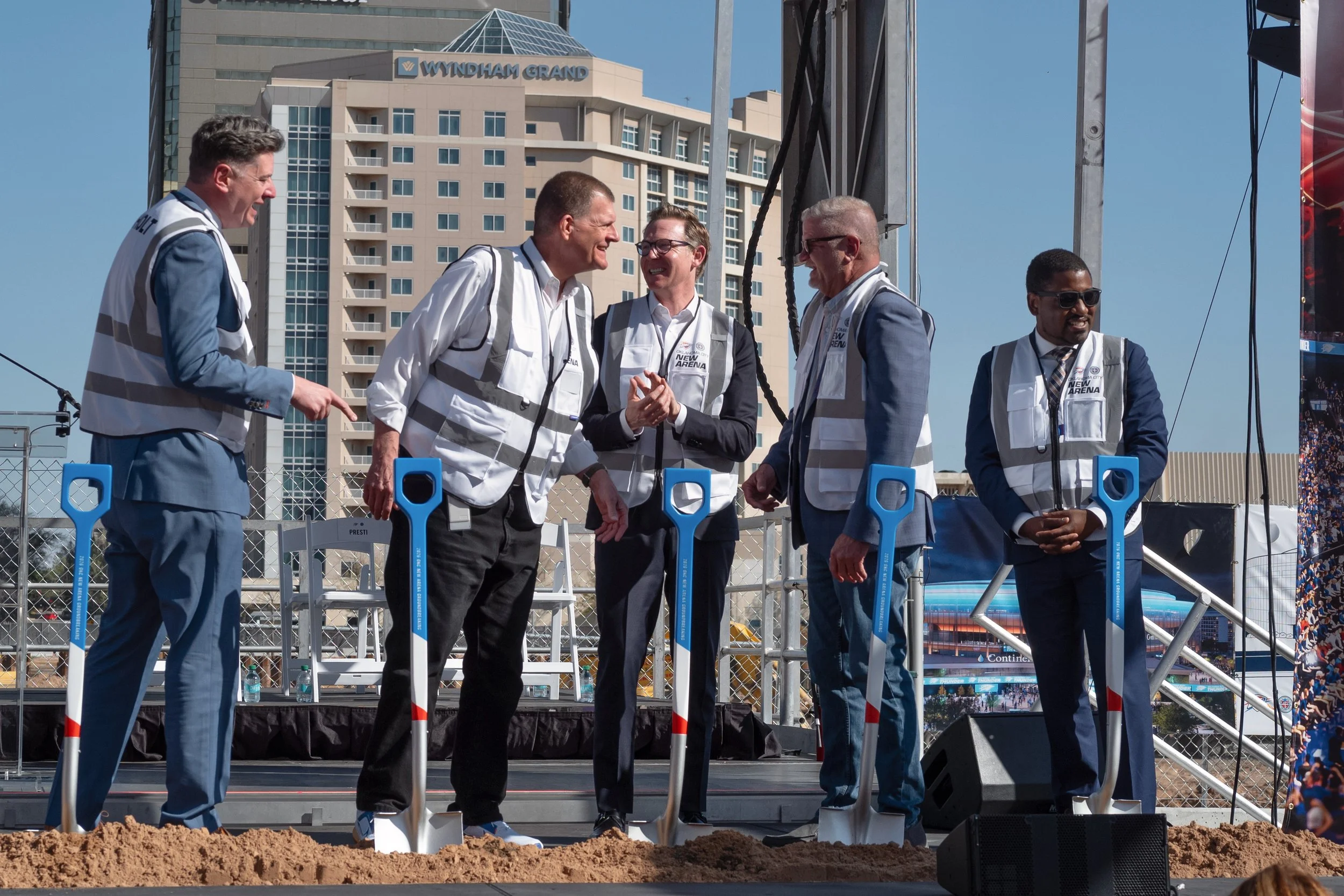 Group of men in suits and vests standing on stage at a groundbreaking ceremony, with shovels in front of them, smiling and talking to each other. Behind them are tall buildings and a clear blue sky.