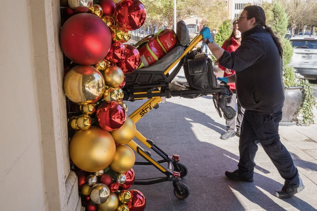A person in black clothing and glasses adjusting a cart with holiday decorations, including red, gold, and silver baubles, on a city sidewalk with trees and parked cars in the background.