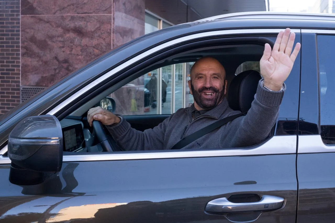 A man with a beard and smiling face sitting in the driver's seat of a car, waving out the window, in an urban area.