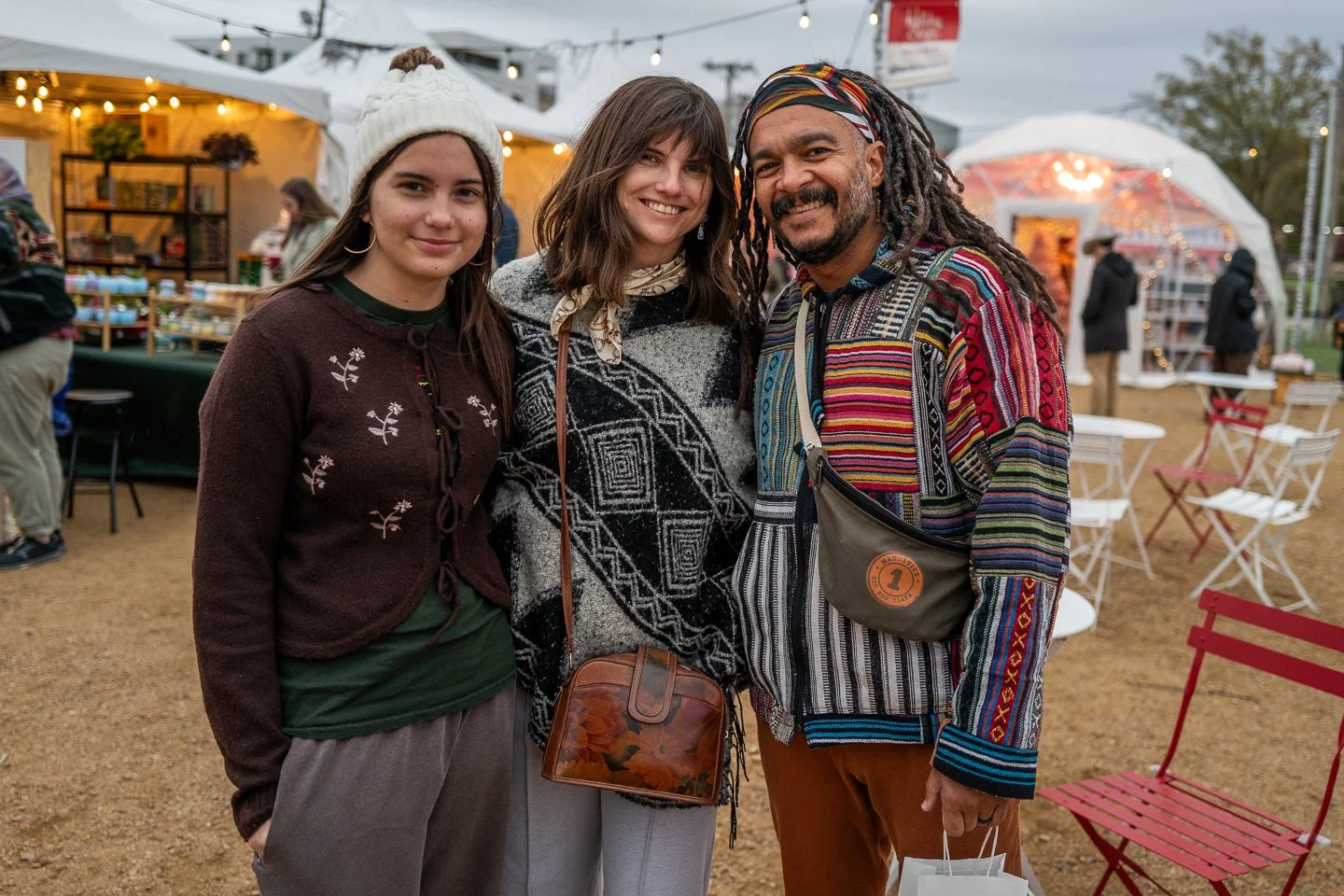 Three people smiling at an outdoor event with tents, string lights, and tables in the background.