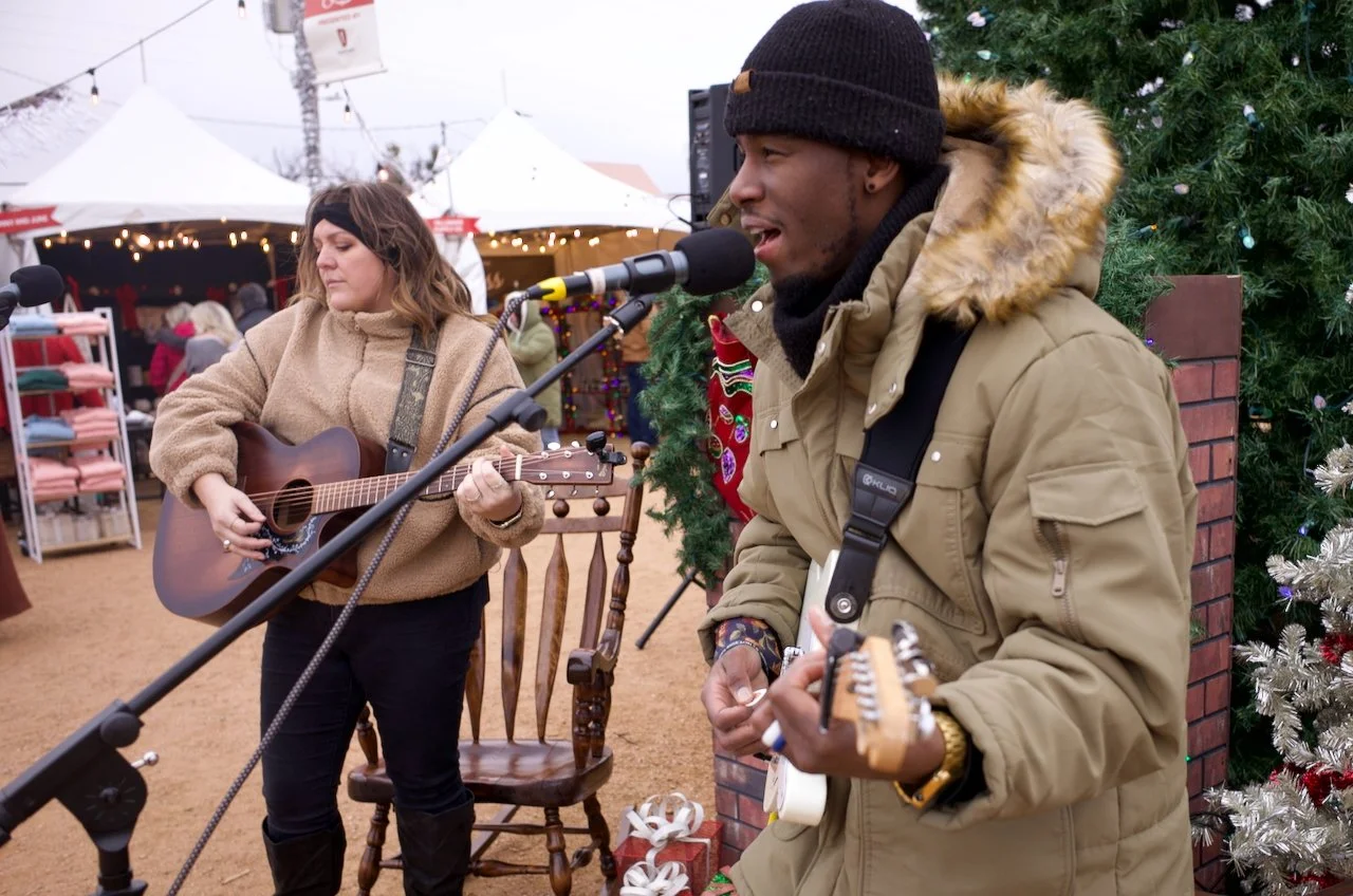 Two musicians performing at an outdoor holiday market, one woman playing an acoustic guitar and one man playing an electric guitar, with Christmas decorations and a Christmas tree in the background.