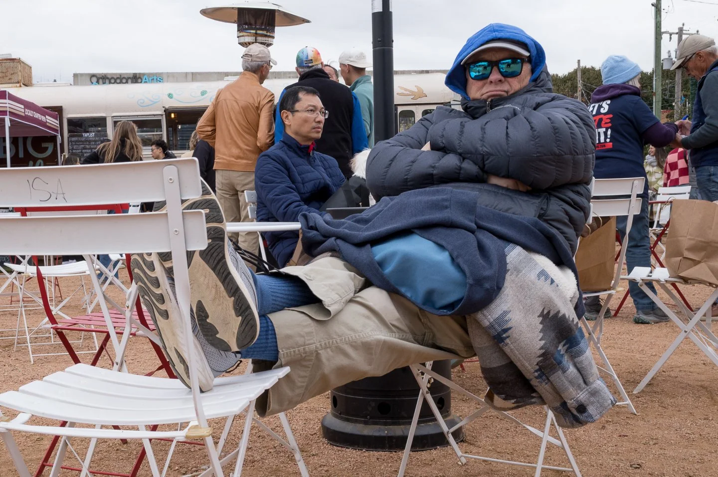 A man sitting with his legs crossed on a white folding chair at an outdoor event, wearing sunglasses, a blue winter jacket, and a cap, with other people standing and chatting in the background.