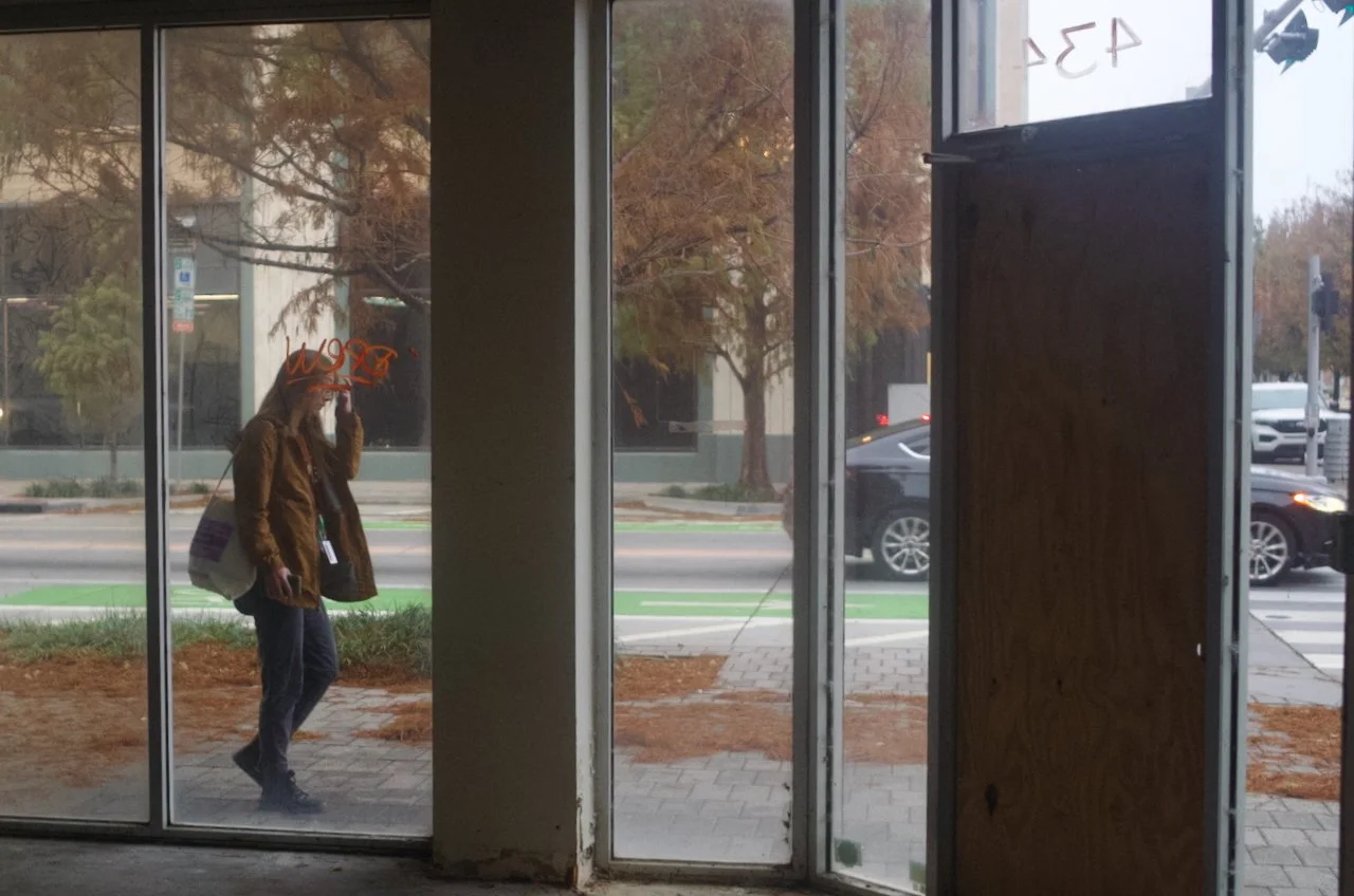 A person is walking outside a building, seen through a large glass window and door. The person is wearing a brown jacket, jeans, and dark shoes, carrying a white bag on their shoulder, and is talking on a cell phone. Trees and cars are visible on the