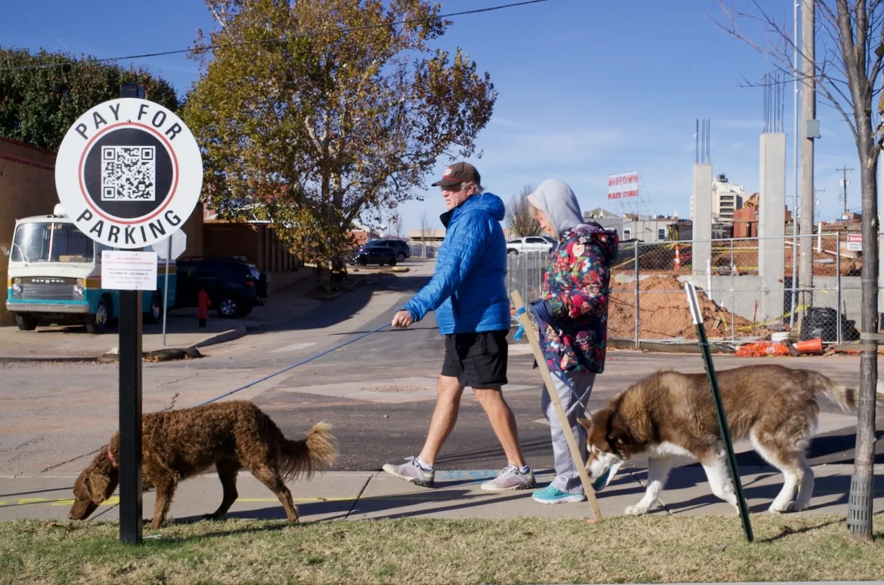 Two people walking dogs on a sidewalk, one with a brown dog and the other with a large brown and white dog, near a sign that says 'Pay for Parking' with a QR code.