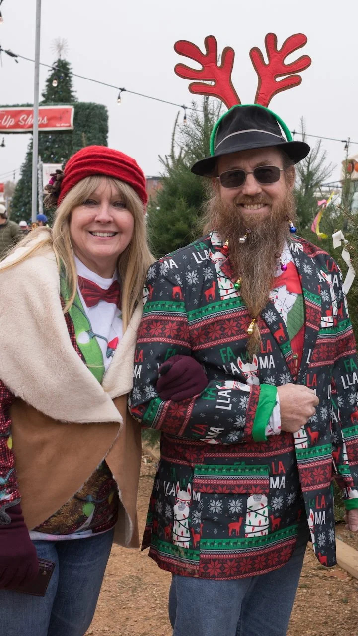 Two people dressed in holiday-themed clothing at an outdoor Christmas market, with Christmas trees and decorations in the background.