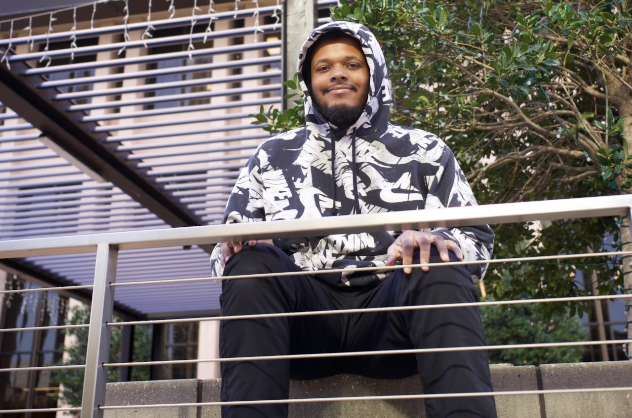 A young man in a black and white patterned hoodie sitting on a ledge outdoors, smiling at the camera, with a metal railing in front of him and trees in the background.