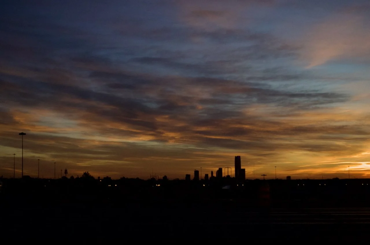 City skyline at sunset with colorful clouds in the sky and silhouetted buildings in the foreground.