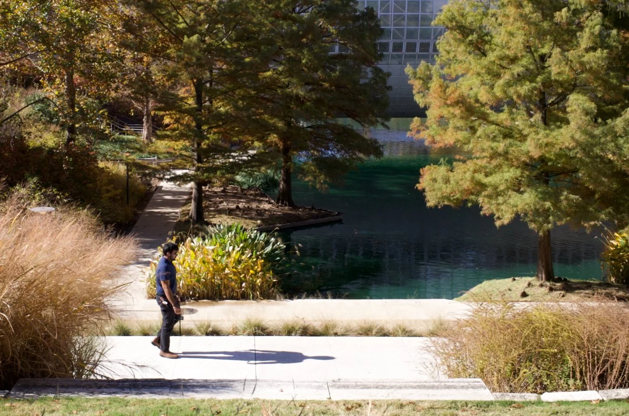 A man walking along a sidewalk near a river or pond, surrounded by trees with fall foliage and ornamental plants.