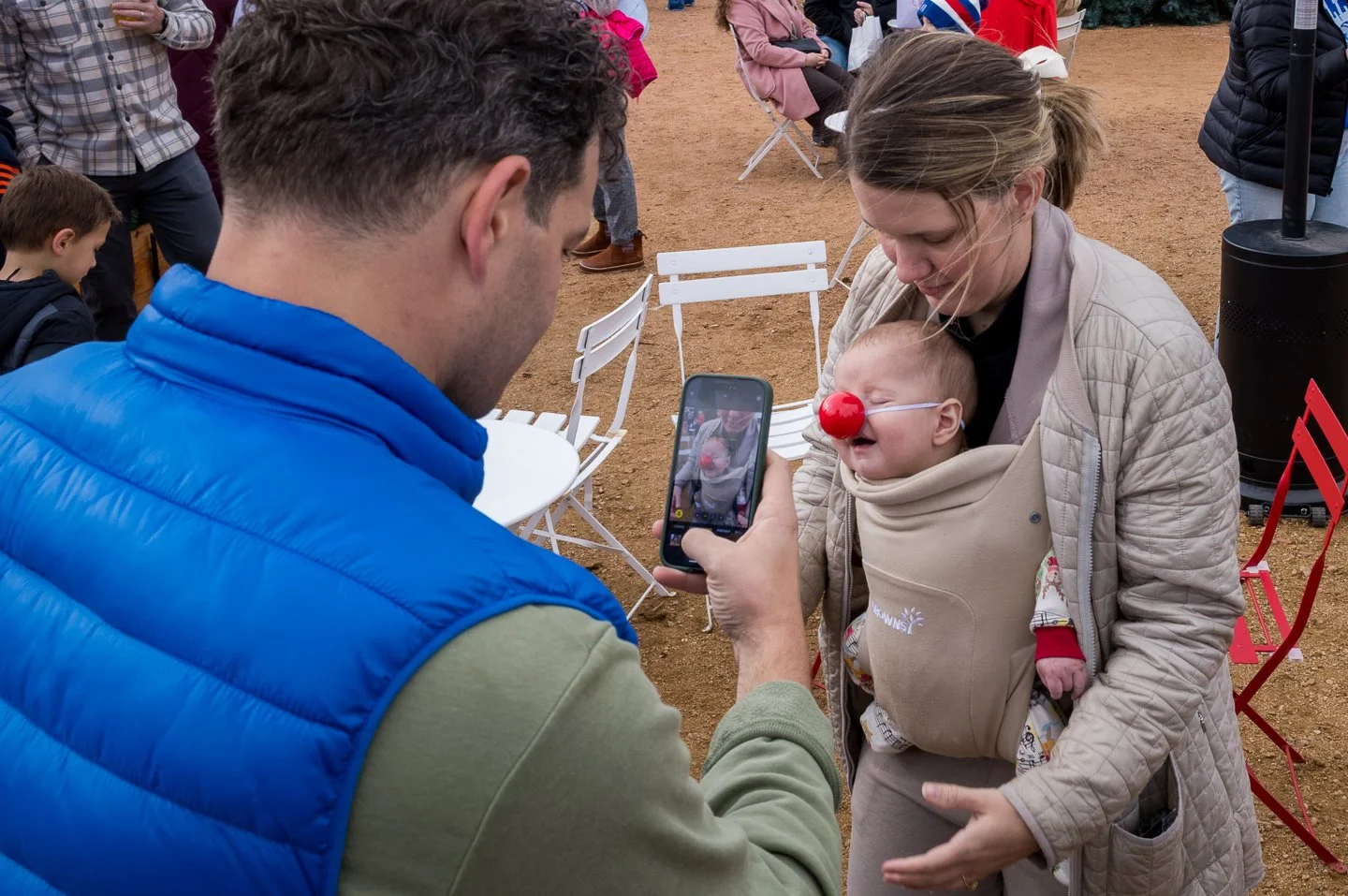 A man takes a photo of a woman holding a baby with a red clown nose at an outdoor event.