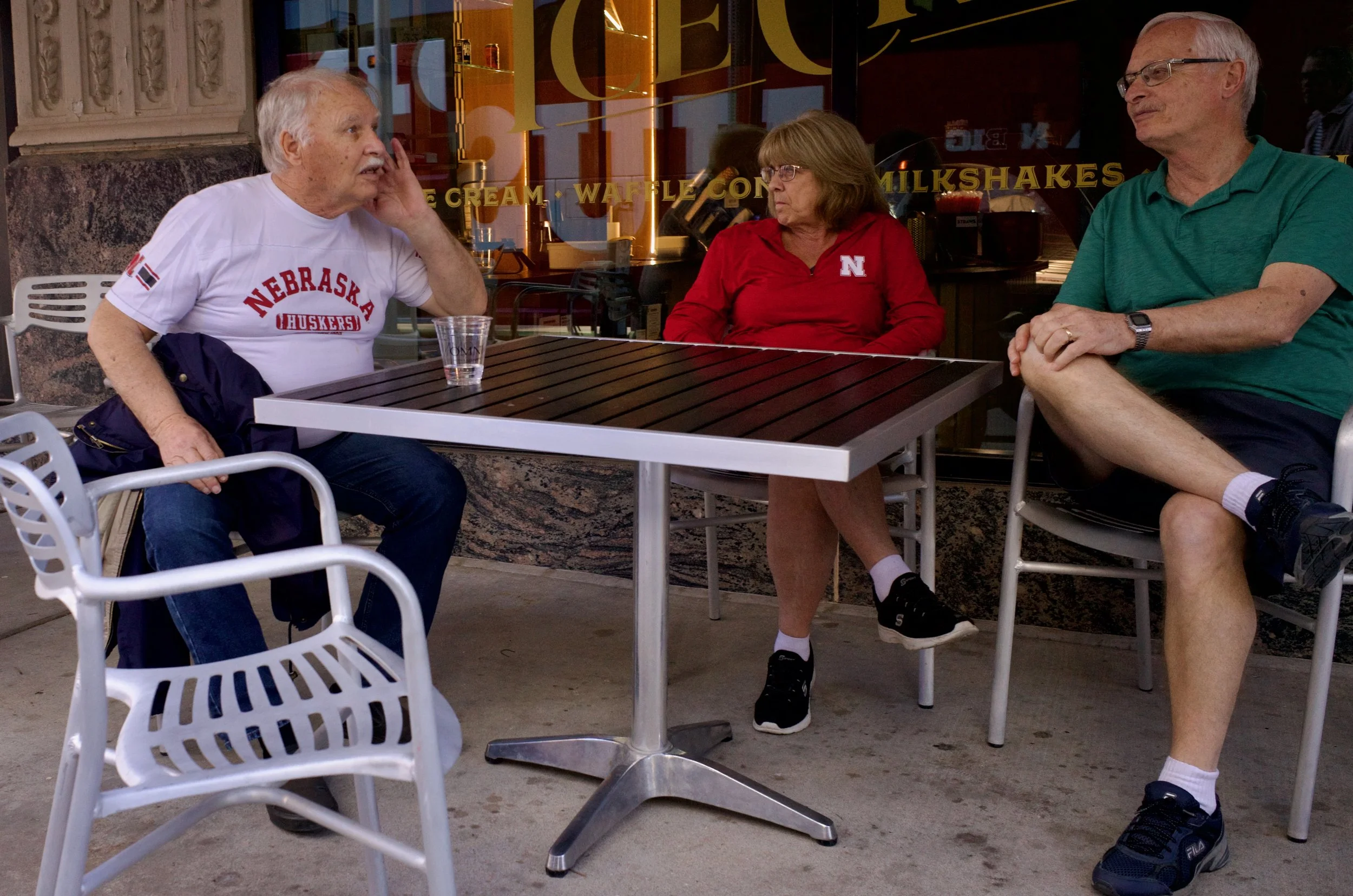Three older adults sitting outdoors at a table in front of a window with decorated lettering. The man on the left is wearing a white Nebraska Huskers T-shirt, the woman in the middle is wearing a red Nebraska jacket, and the man on the right is weari