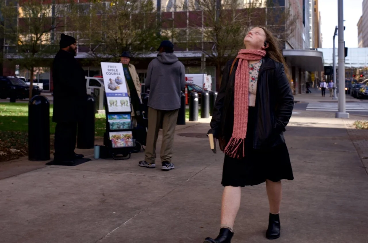 A woman in downtown OKC with long hair, wearing a black jacket, scarf, and skirt, stands on a city sidewalk looking upward. In the background, three men are near an outdoor stand with a sign that reads "Free Bible Course."