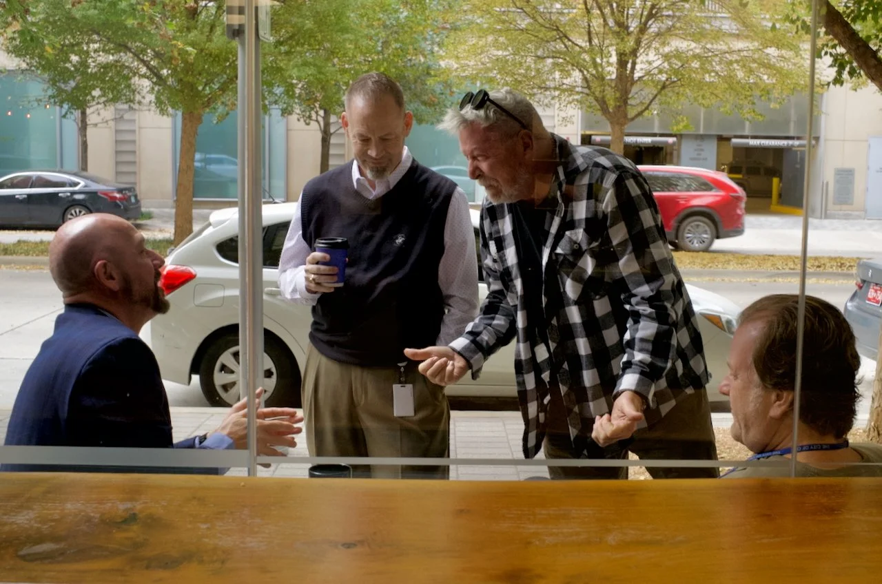 Four men having a lively discussion outside a cafe or restaurant, with cars parked on the street behind them. Three men are standing, one man is sitting inside. The scene takes place during daytime, with autumn-colored trees visible in the background