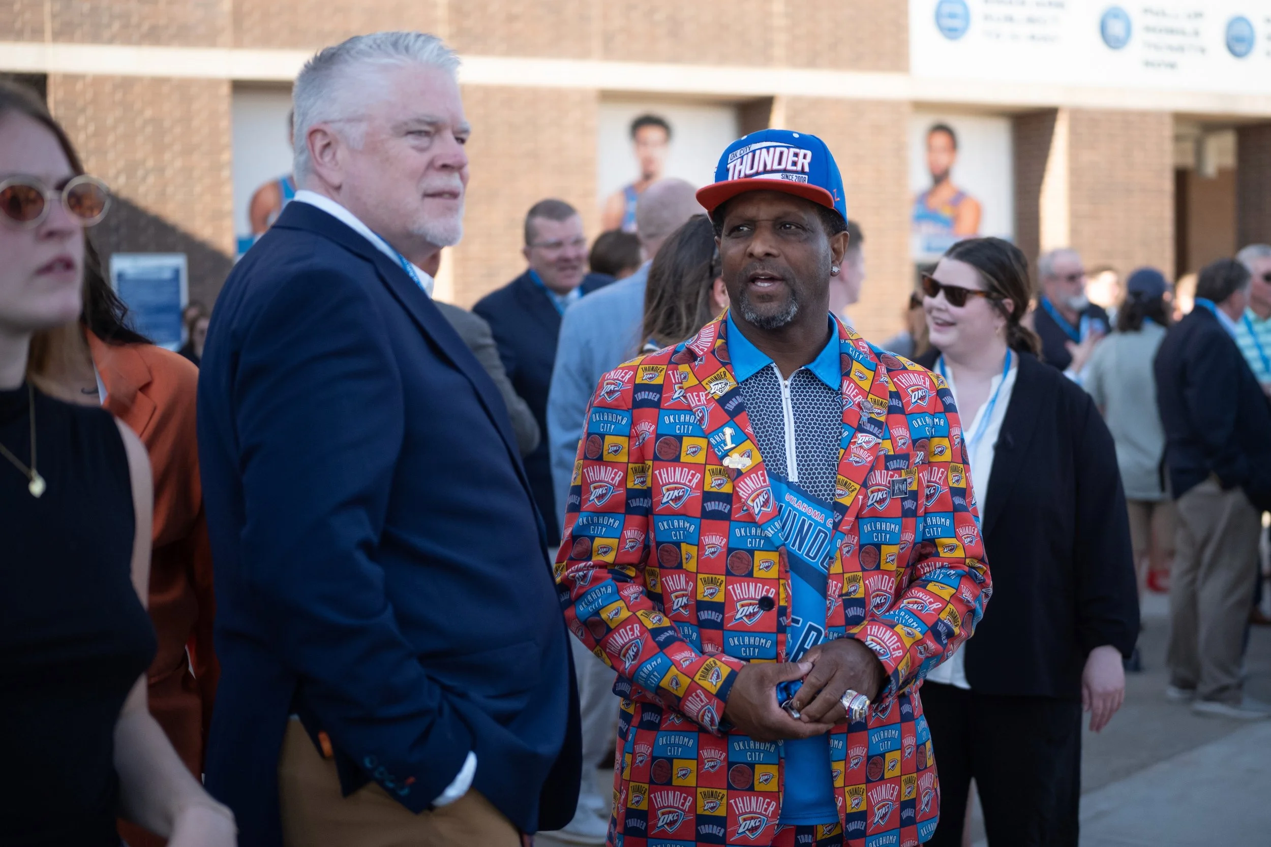 A man wearing a colorful Oklahoma City Thunder jacket and cap is talking to an older man in a blue suit at an outdoor event, with people and posters in the background.