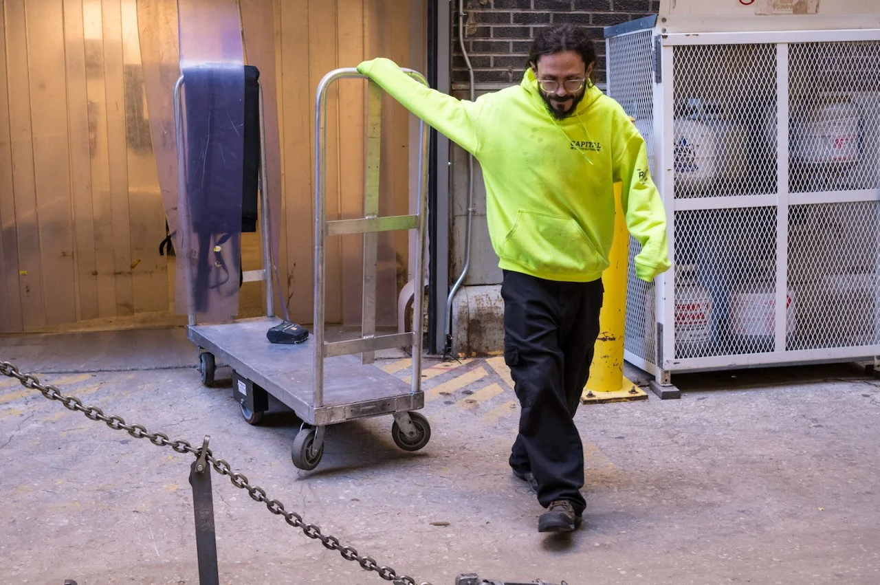 Man with glasses and a beard wearing a bright yellow hoodie and dark pants walking past a metal cart on an outdoor pavement.