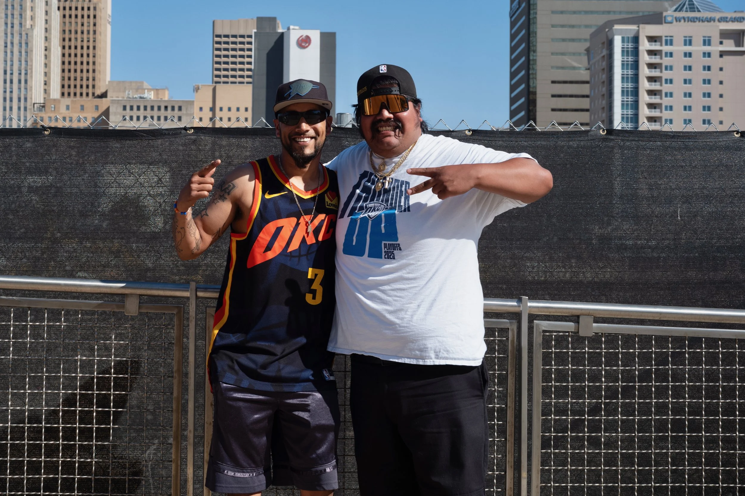 Two men posing together outdoors with city buildings in the background. The man on the left is wearing a black Oklahoma City Thunder basketball jersey, sunglasses, and a cap. The man on the right is wearing a white T-shirt, large sunglasses, and a ca