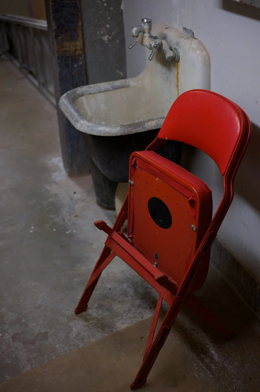 An old, weathered utility sink attached to a wall with a rusty drain and faucet, next to a red folding chair with a circular cutout on the backrest in an industrial or workshop setting.