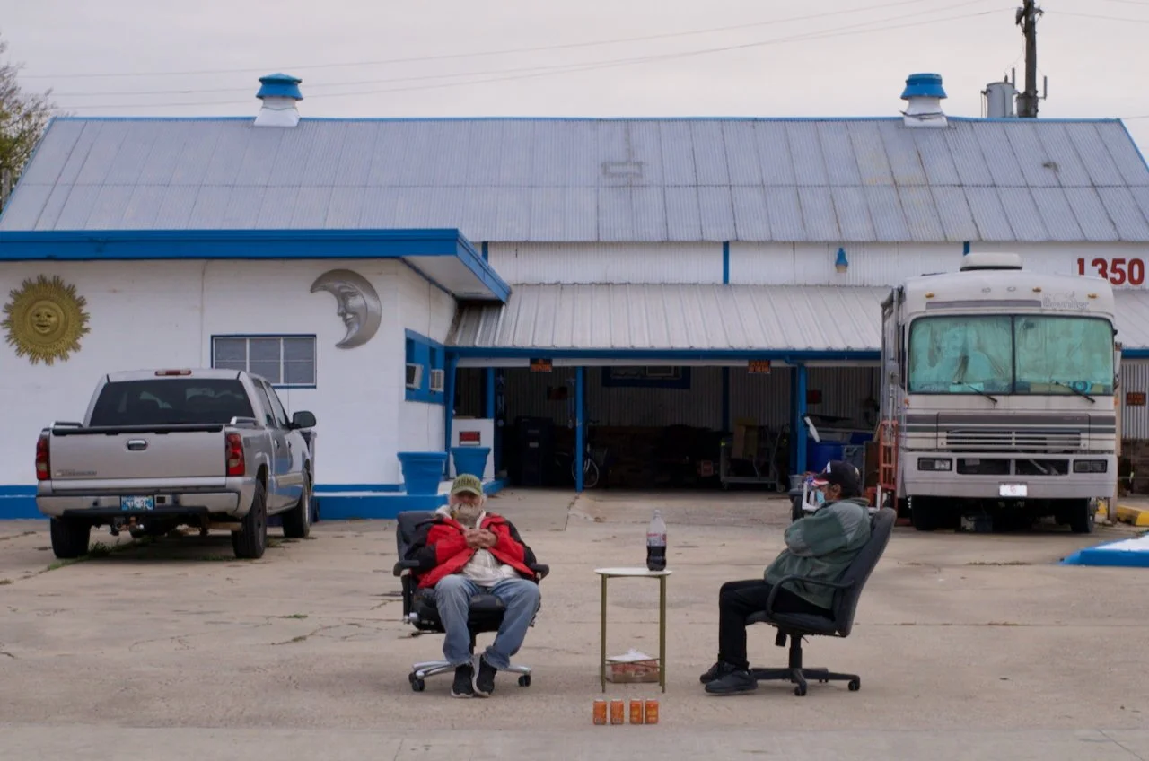 Two men sit outdoors in front of a building with a painted moon and sun, drinking soda. One is wearing a red jacket and the other a gray hoodie. There is a small table with a bottle of soda on it, and several orange cans on the ground. A truck and a 