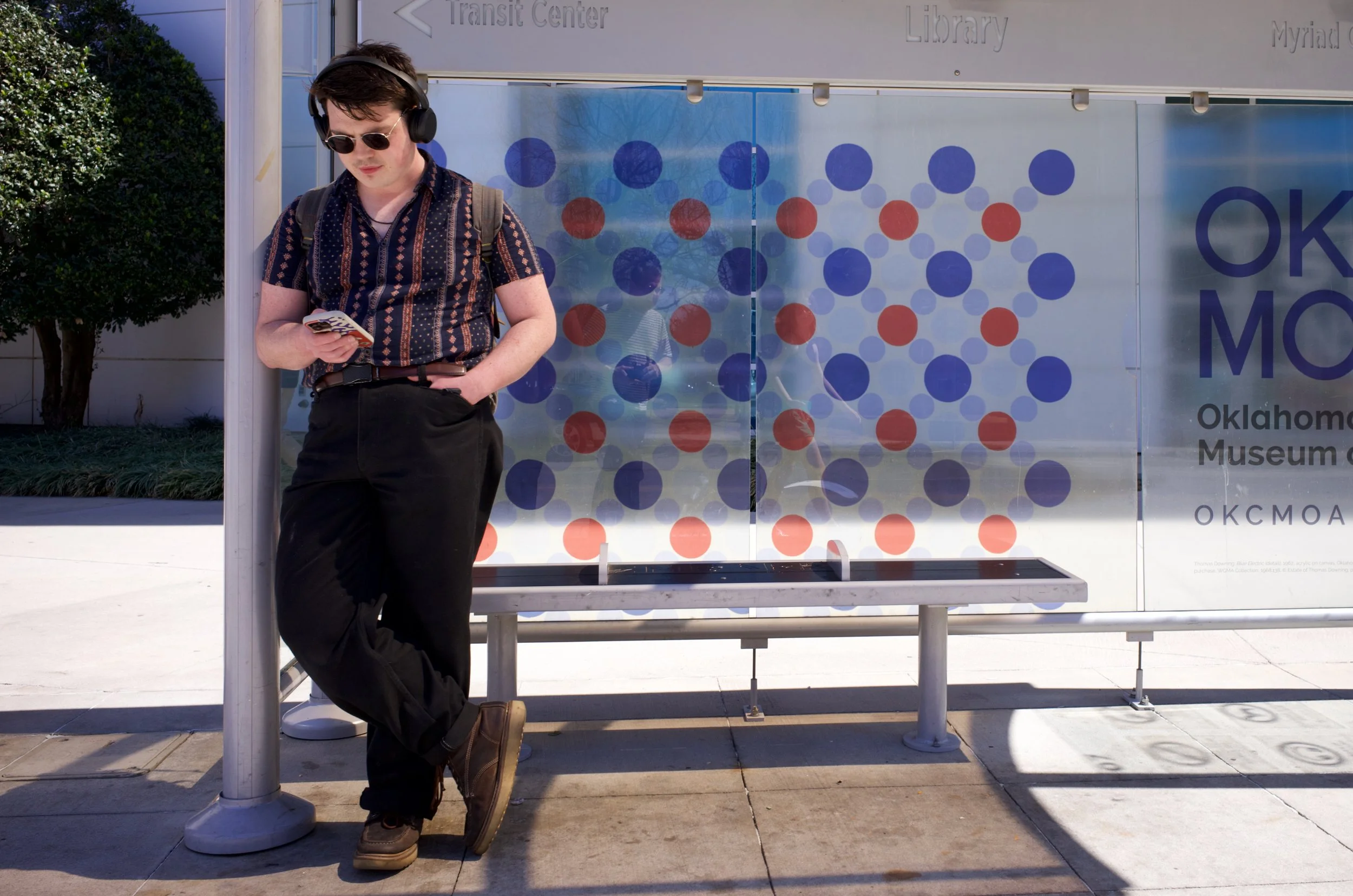 A young man with headphones, sunglasses, and a backpack leaning against a pole at a bus stop, looking down at his phone. Behind him is a sign for the Oklahoma Museum of Science and History, with a patterned window and some trees visible.