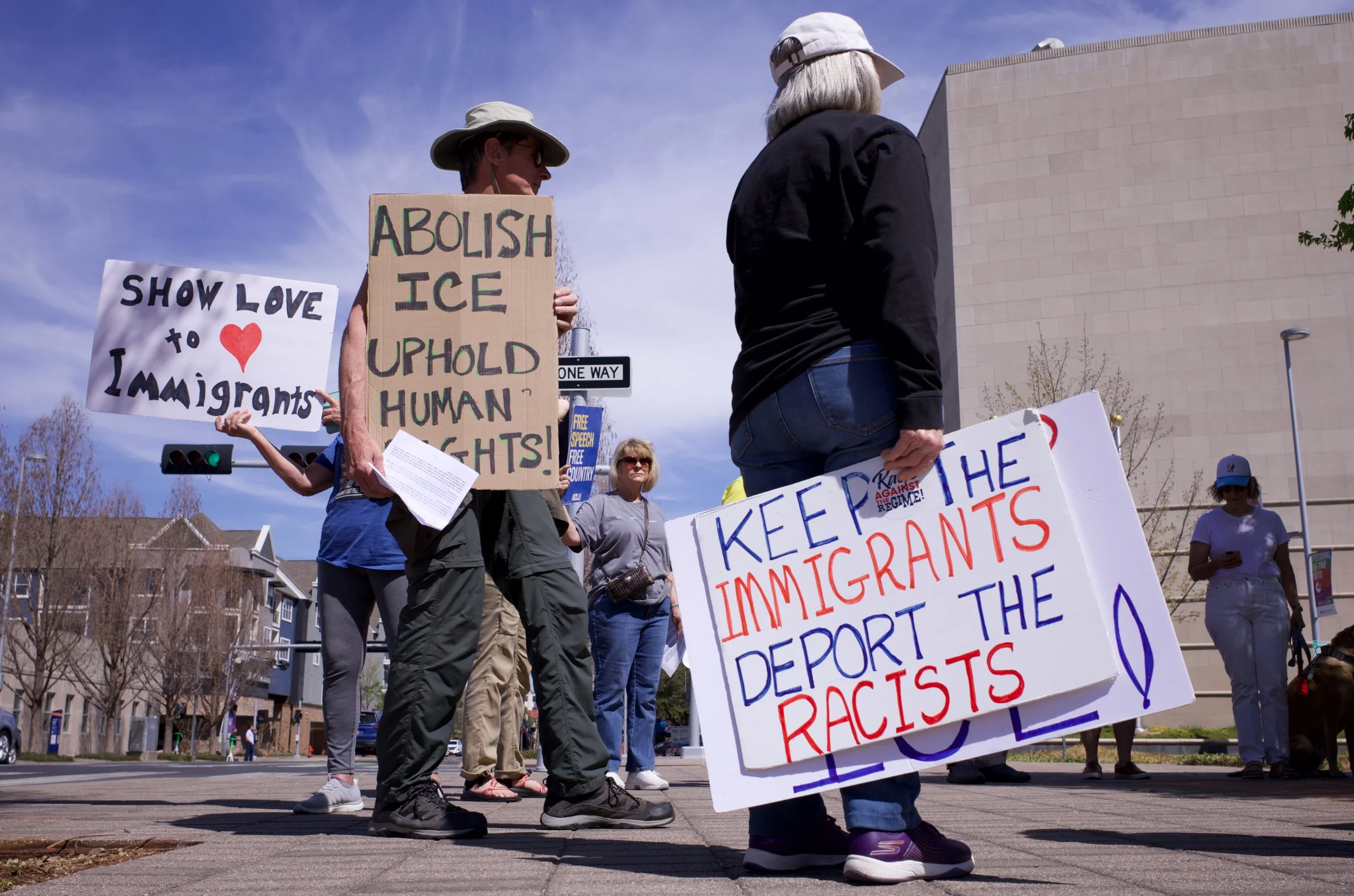 Group of people protesting for immigrant rights, holding signs with messages such as "Show love to immigrants," "Abolish ICE," and "Keep the immigrants out the racists!" on a street with buildings and trees in the background.