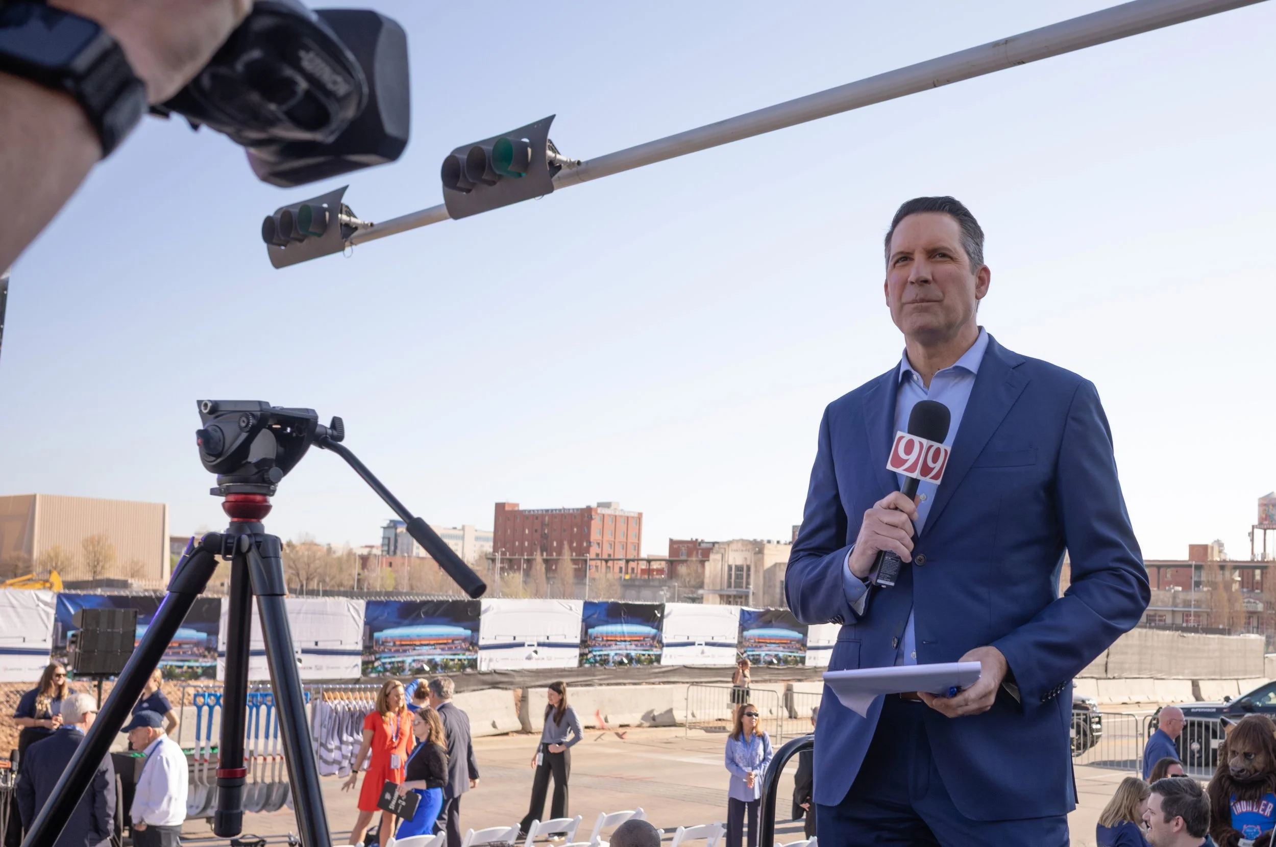A male news reporter in a blue suit holding a microphone with the '9' logo stands outdoors during a press event, with a camera on a tripod filming him. Several people are seen in the background at an open area under a clear sky.