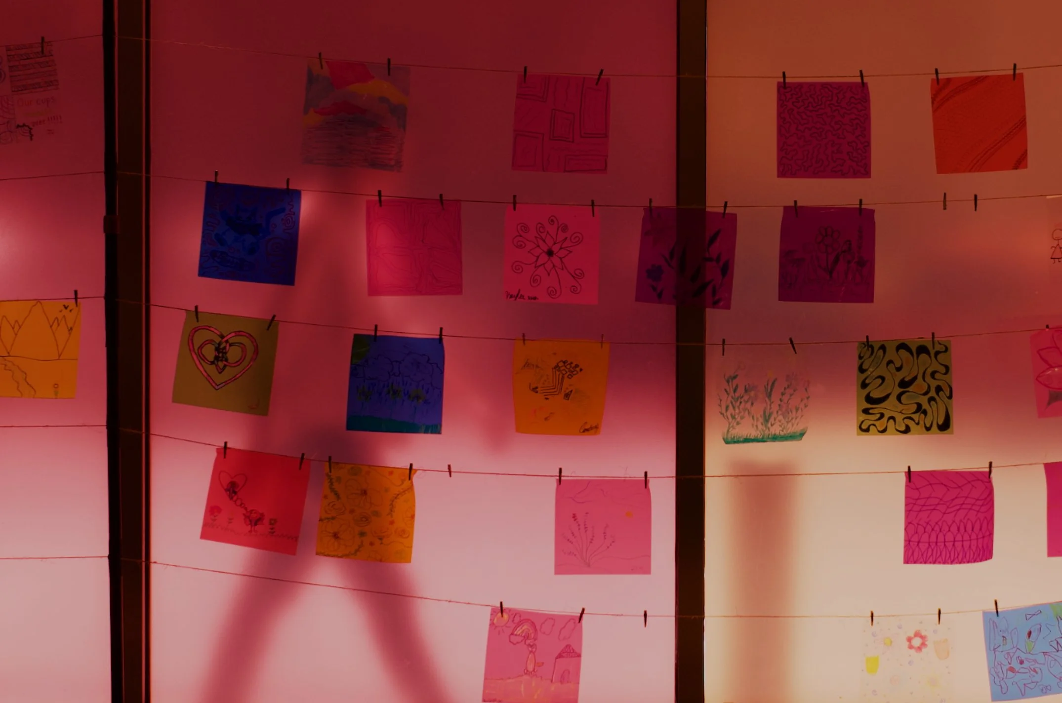Colorful drawings and artwork hanging on thin strings with clothespins, illuminated by red and pink lighting.