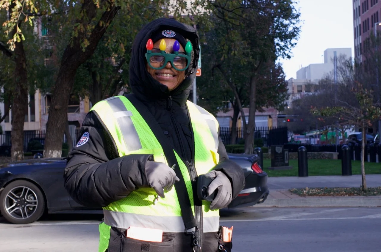 A person dressed as a traffic officer wearing a neon yellow reflective vest, winter coat, gloves, and a black hoodie, standing on a city street with trees and parked cars in the background. The person is smiling and wearing festive glasses and a colo