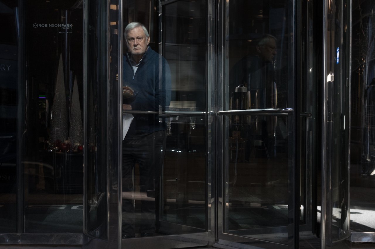 Older man with gray hair and a serious expression standing inside a revolving glass door, holding a small object, possibly a snack or food item.