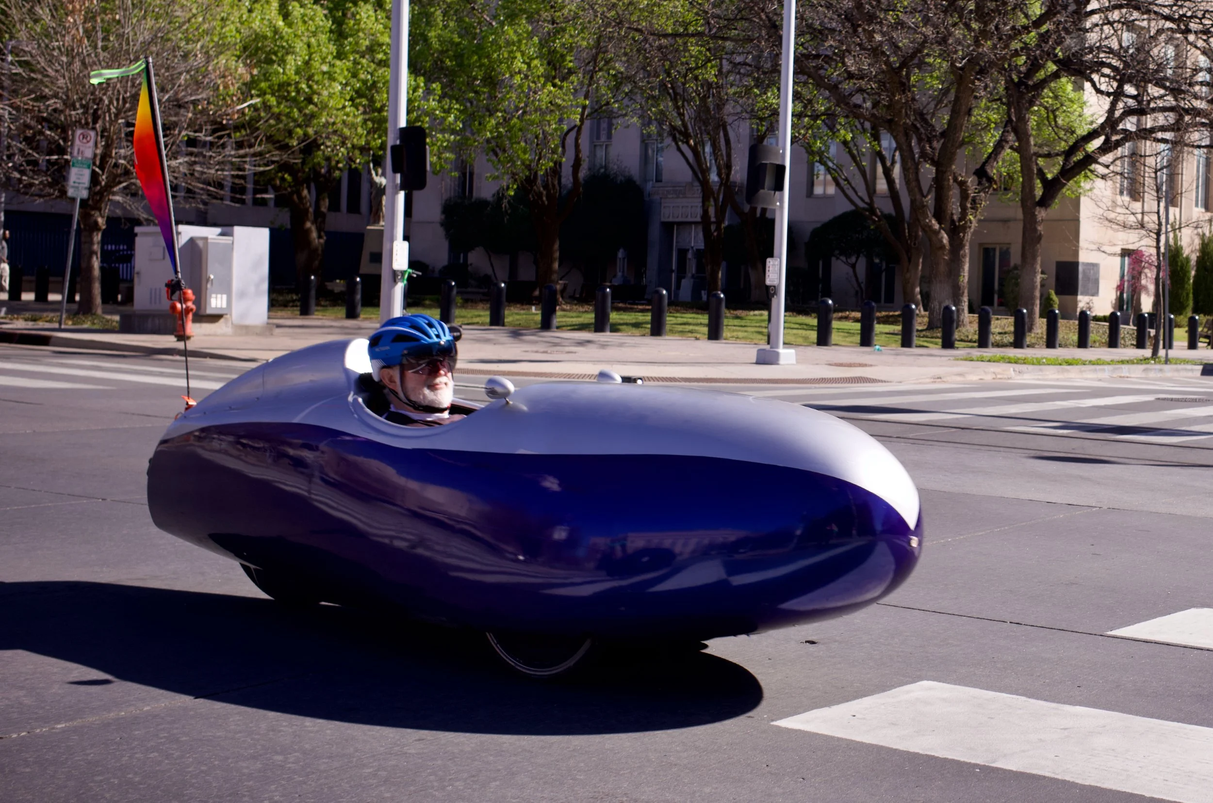 A man wearing a blue helmet riding a futuristic, aerodynamic velomobile with a rainbow flag at the back, on an urban street with trees and buildings in the background.