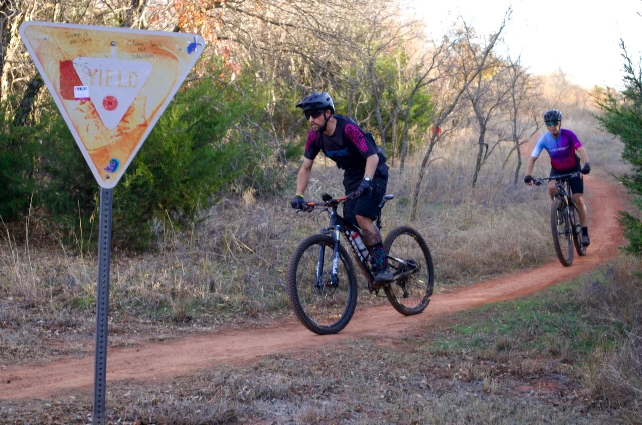 Two mountain bikers riding on a dirt trail through a wooded area with sparse trees and bushes, near a yellowed yield sign with handwritten notes.
