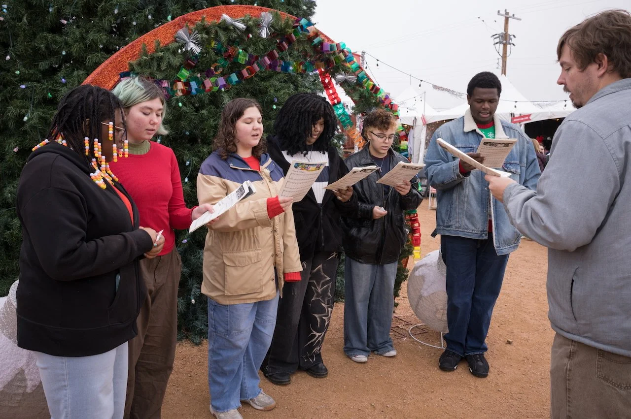 Group of six young people singing or reading from song sheets outdoors during a festive event, standing in front of a decorated Christmas tree with garlands and ornaments.