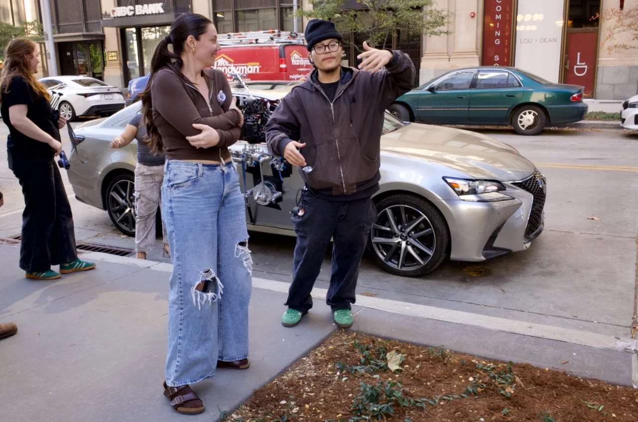 A group of people on a city sidewalk, with a woman in ripped jeans and a man in a hoodie talking, and a silver Lexus parked behind them. Other pedestrians are nearby, and a camera rig is visible behind the woman.