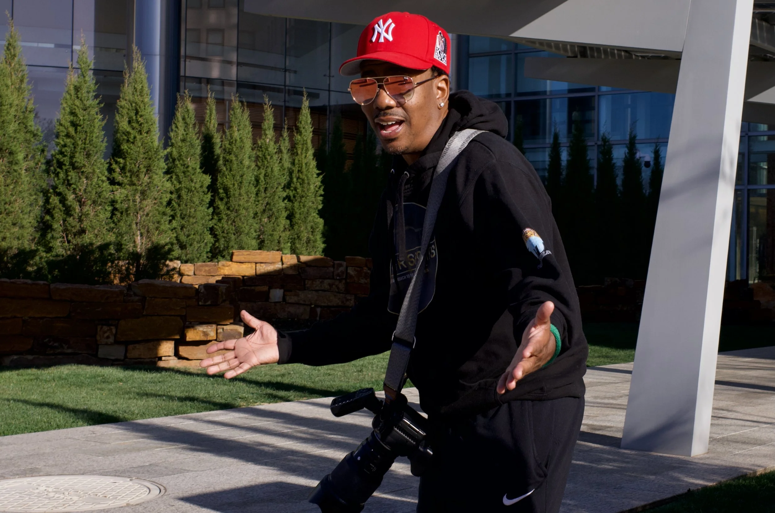 Man wearing a red cap, sunglasses, and a black hoodie with a camera around his neck, gesturing with open hands outdoors in front of a modern building and green trees.