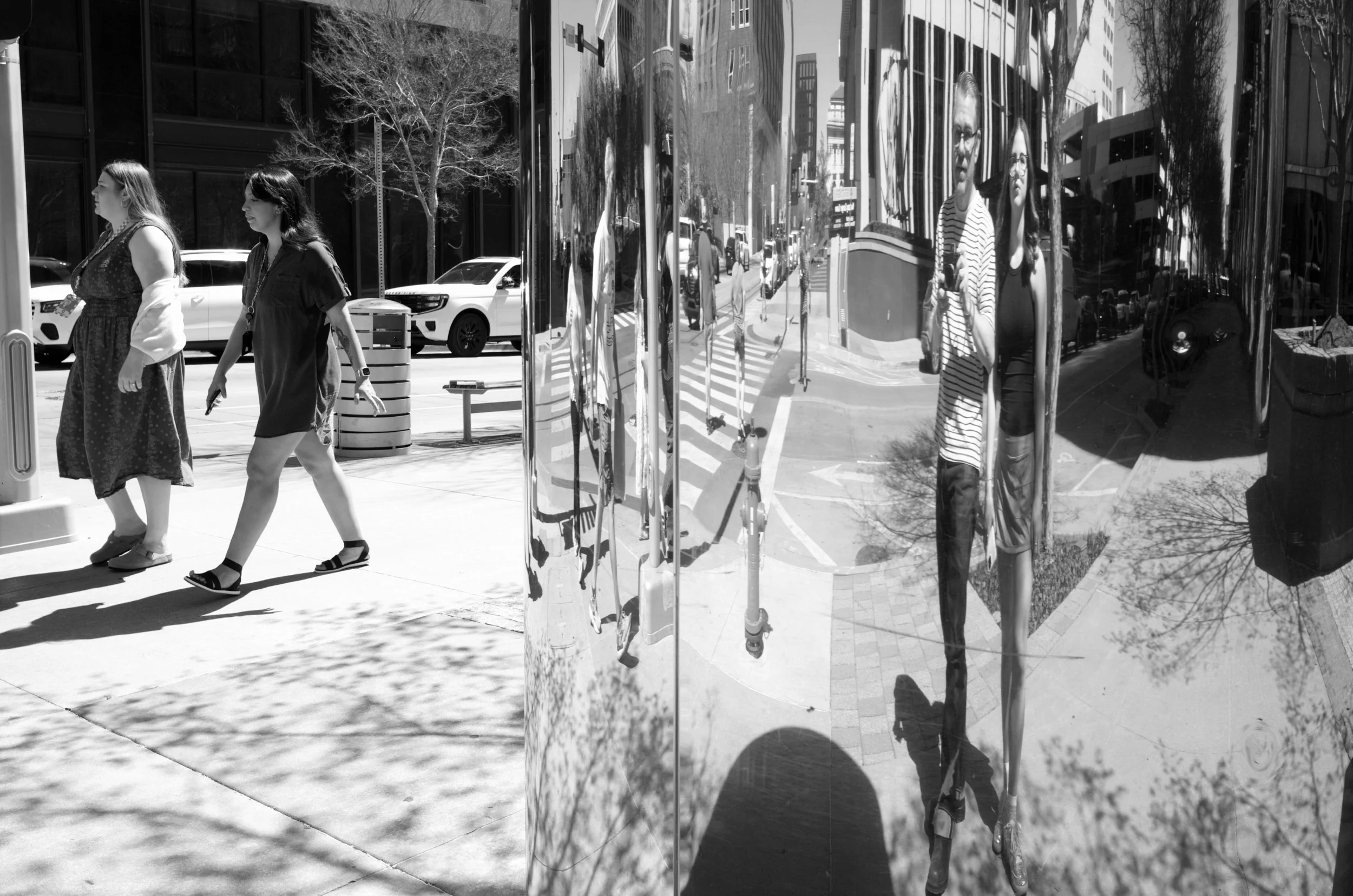 A black and white photo of city street with pedestrians, parked cars, reflected buildings, and two mannequins in striped clothing reflected in a shiny surface.