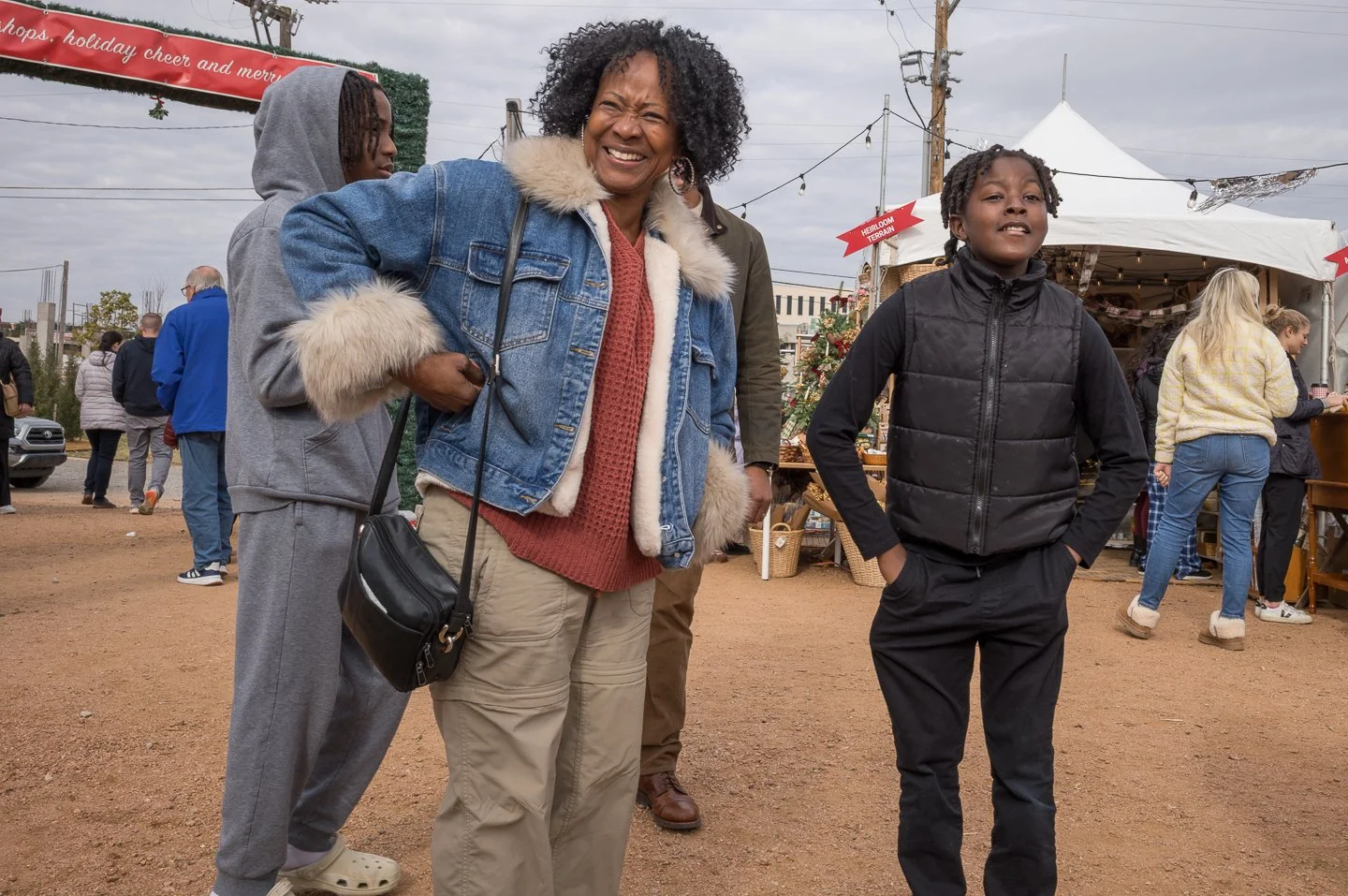 A group of people at an outdoor market, including a smiling woman in a denim jacket and a girl in a black jacket, with other market-goers and stalls visible in the background.