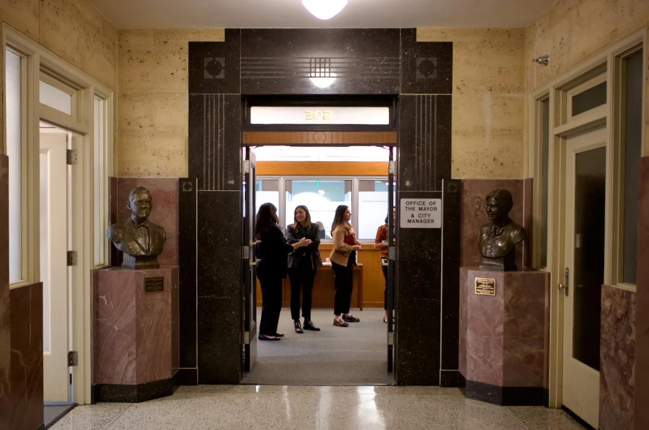 View through the entrance of a government building with bust statues on pedestals on either side, a sign for the Office of the Mayor and City Manager, and people inside engaged in conversation.
