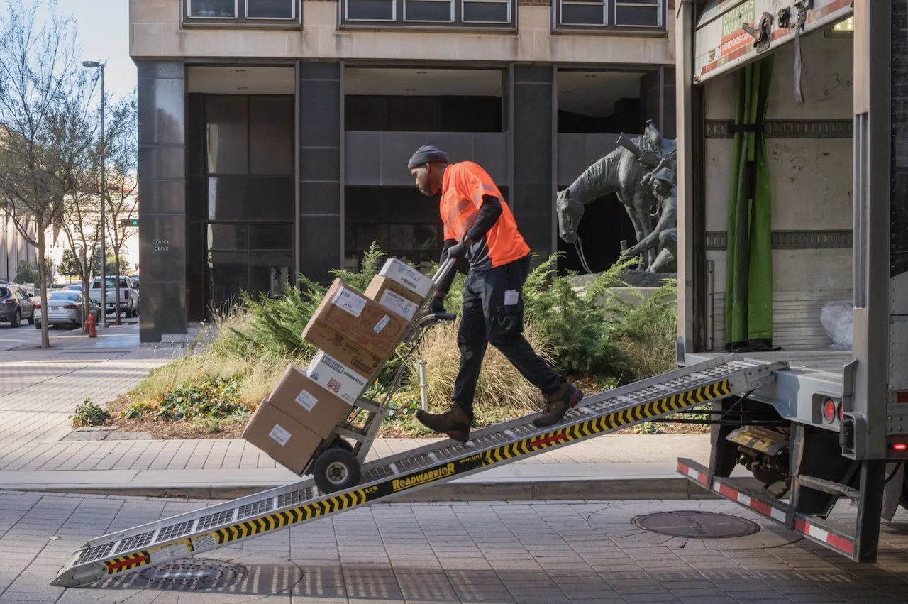 A man in a bright orange safety vest unloads boxes from a delivery truck on a ramp in an urban area with trees and parked cars, and a building with a sculpture of horses in the background.