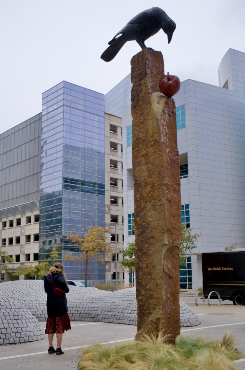 A woman standing in a downtown OKC urban plaza in front of a tall sculpture of a bird perched on a large stone pillar with an apple carved into it, surrounded by modern glass buildings.
