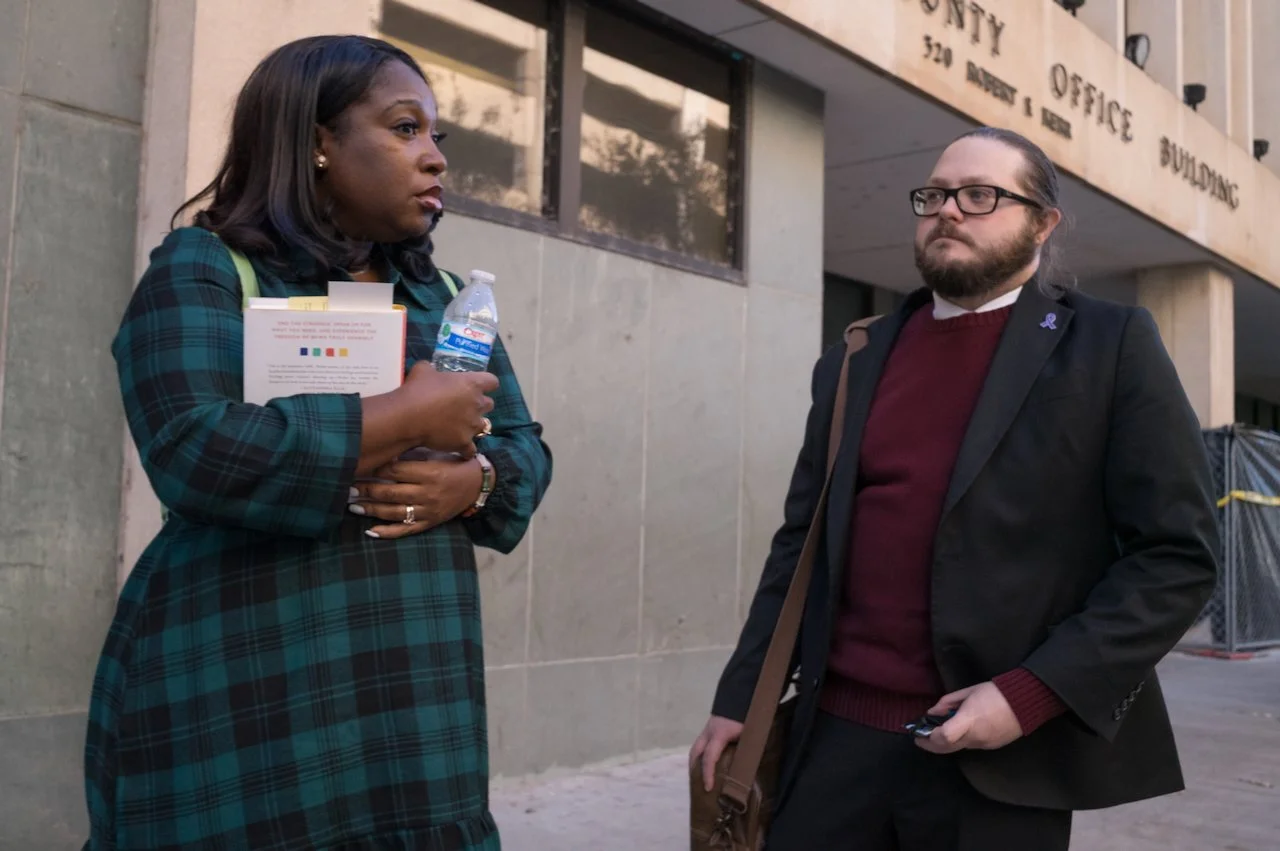Two people standing outside a building engaged in conversation. One woman in a plaid dress holding a water bottle and notebooks, the other man with glasses and a beard holding a phone, both appearing serious.