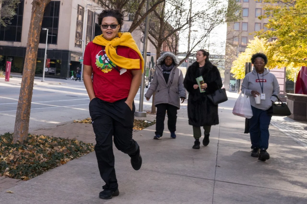 Four women walking on a city sidewalk with trees and buildings in the background, one wearing a red shirt with a colorful graphic, sunglasses, and a yellow jacket draped over her shoulders, smiling and leading the group.