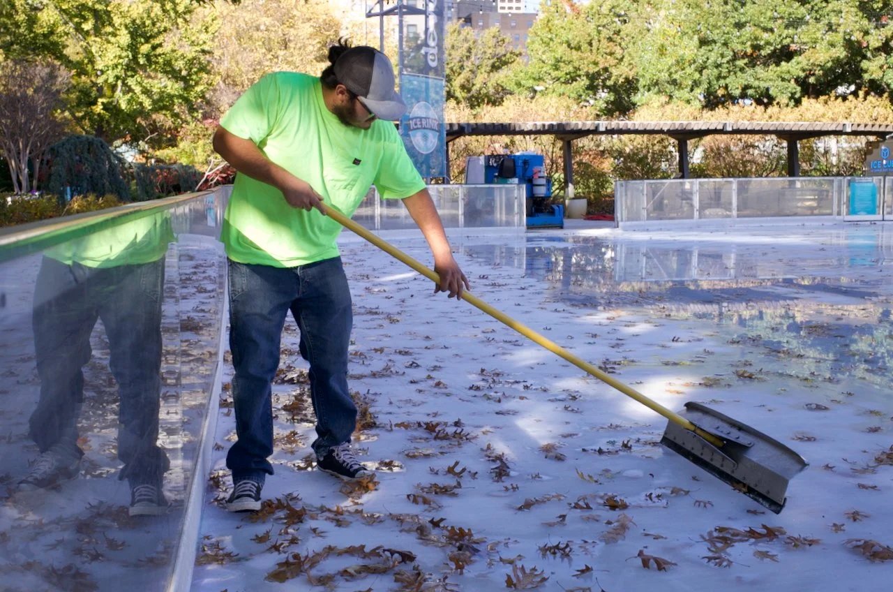 A person wearing a neon green shirt, jeans, and a cap is sweeping fallen leaves off the ice at an outdoor ice skating rink.