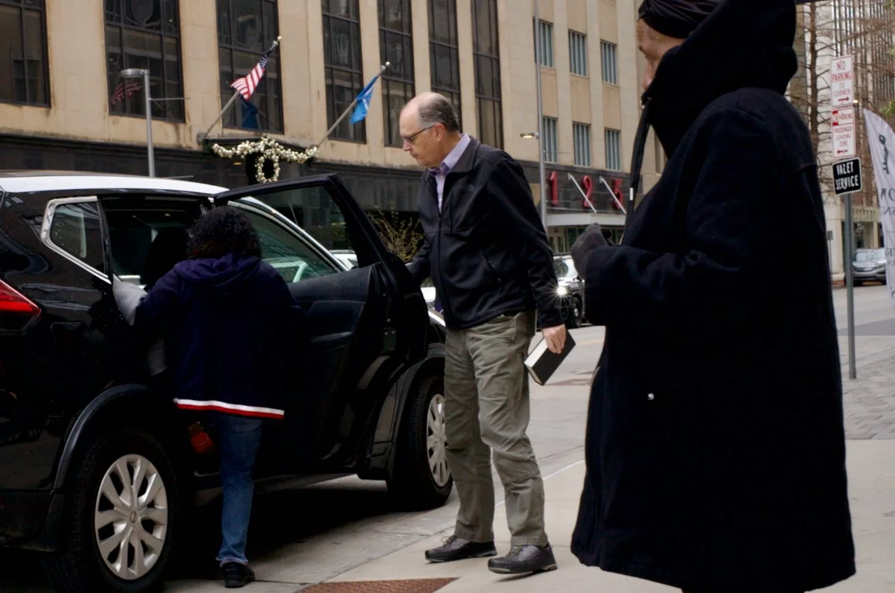 Three people, two adults and one child, are outside on a city street near a black SUV. The woman is getting into the back seat, the man is holding a book, and the third person is dressed in a dark coat and hoodie.