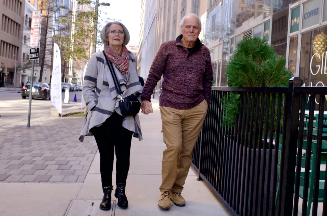 An elderly couple walking hand in hand on a city sidewalk, with tall buildings and storefronts in the background.